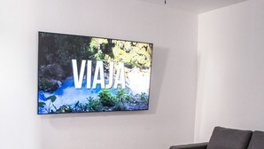 Una sala de estar moderna con un sofá modular, una mesita de centro con objetos decorativos y un televisor grande montado en la pared mostrando 'VIAJA'.