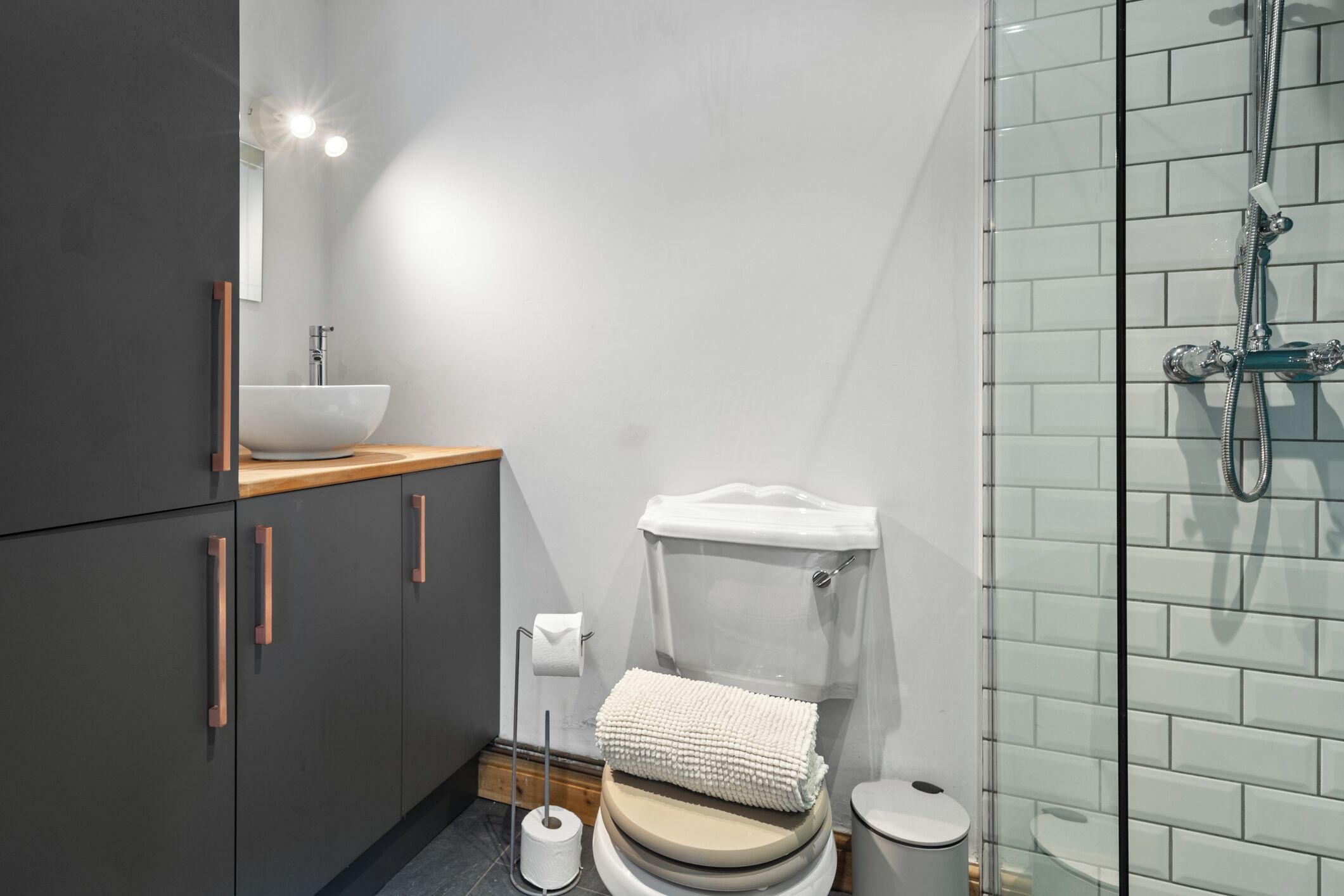 Un baño moderno con inodoro blanco, tocador de madera, cabina de ducha con mampara de vidrio y lavamanos blanco.