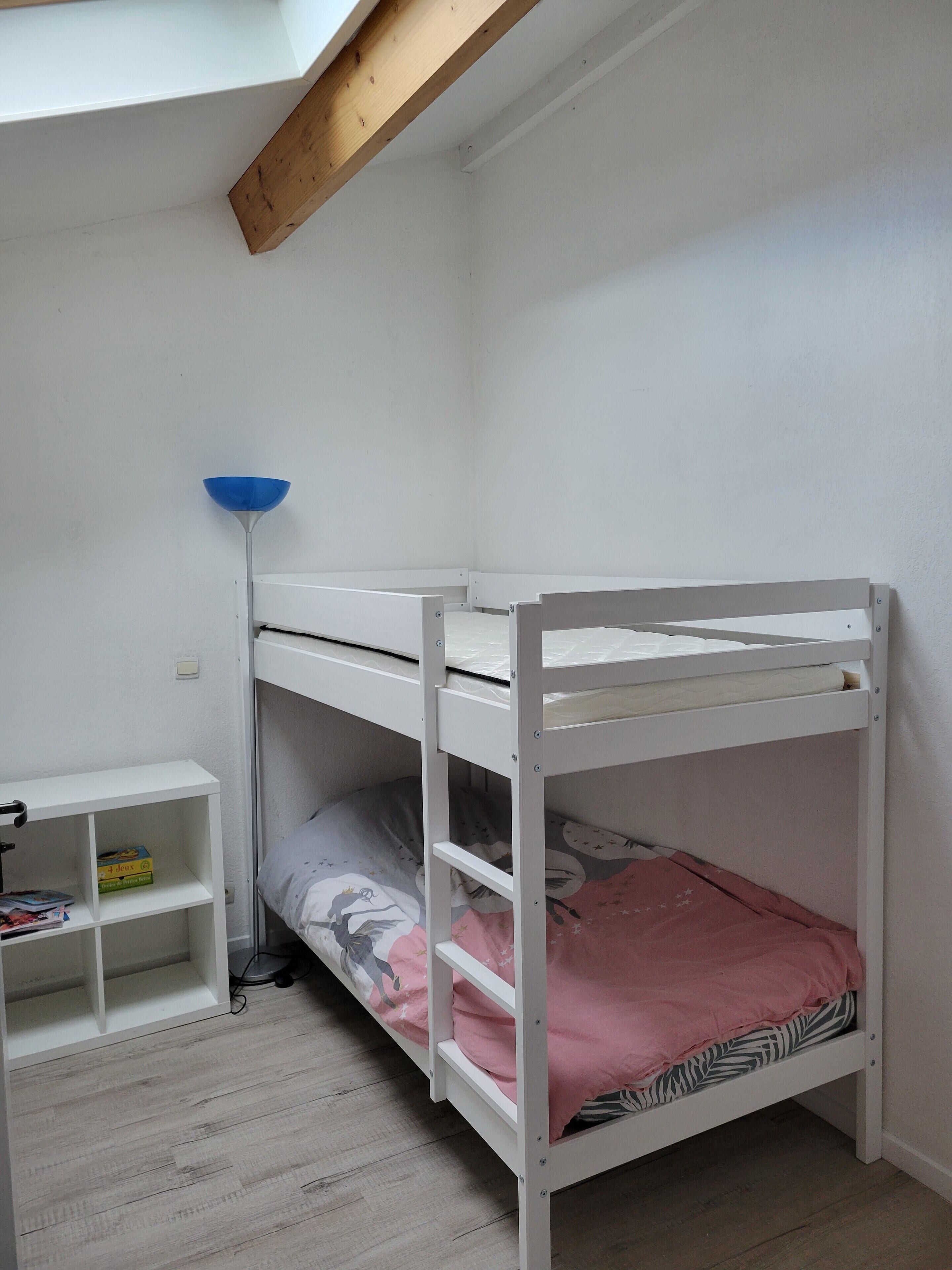 A bunk bed with a lower bunk, a white shelf unit, and a blue floor lamp in a room with white walls and wooden beams.