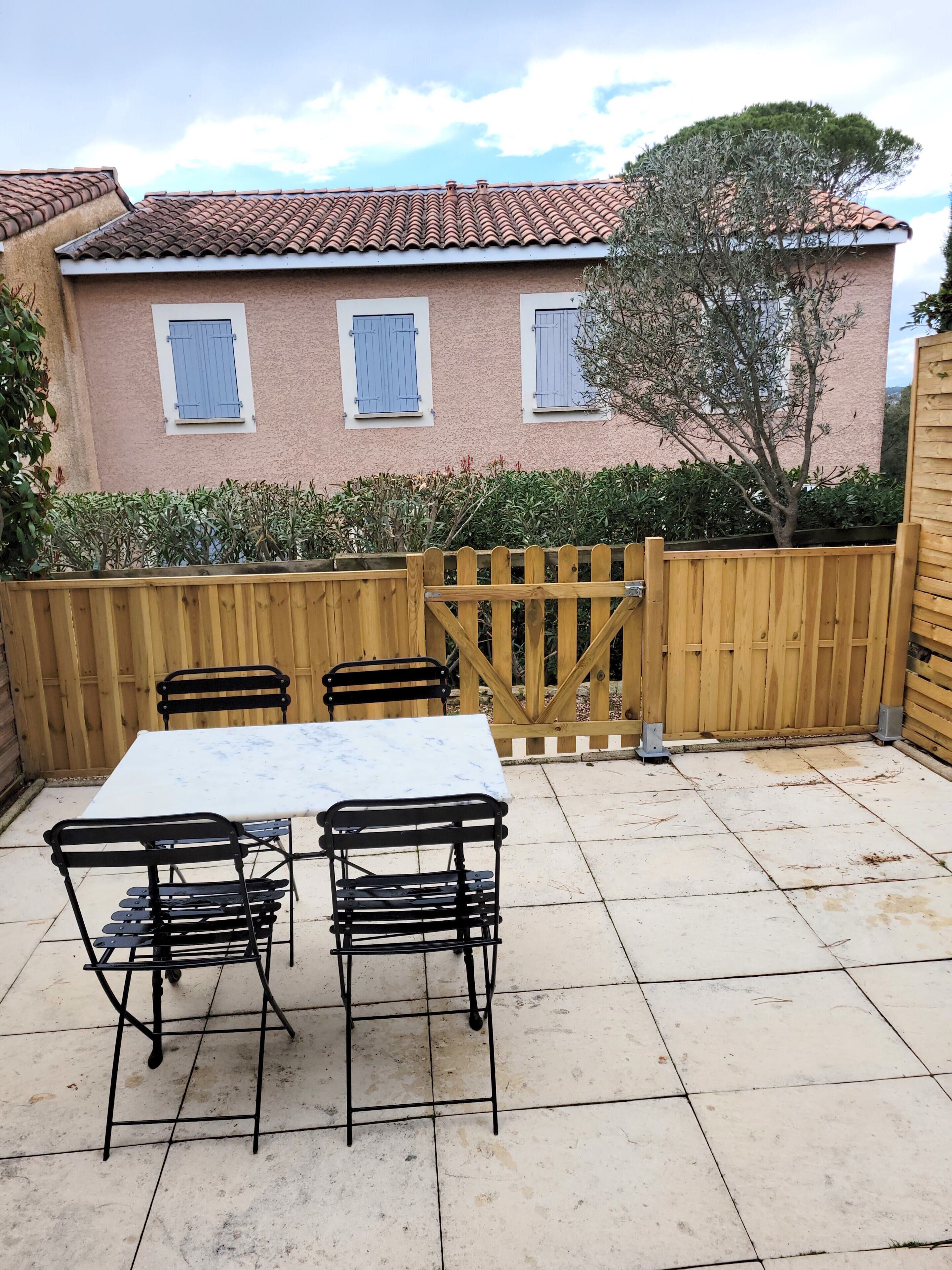 Eine Terrasse mit Tisch und Stühlen, ein Holzzaun und ein Gebäude mit Ziegeldach im Hintergrund.