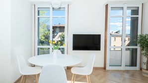 A modern living room with a white round table, four white chairs, a flat-screen TV mounted on the wall, and large windows with a view of buildings and trees. - Appartamento Volta 4 (Cernobbio)