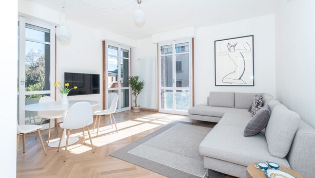 A modern living room with a grey sofa, white chairs, a dining table, and a TV. - Appartamento Volta 4 (Cernobbio)