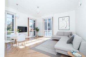 A modern living room with a grey sofa, white chairs, a dining table, and a TV. - Appartamento Volta 4 (Cernobbio)