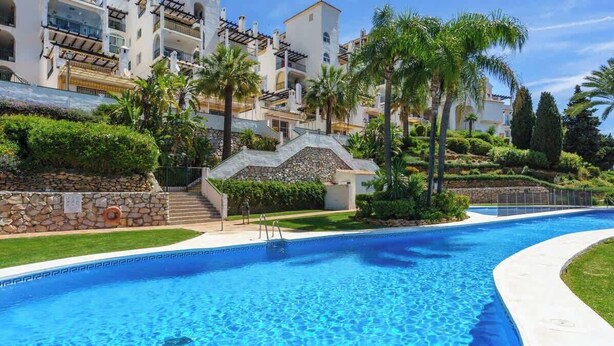 A swimming pool with a stone wall and palm trees, surrounded by multi-story buildings with balconies. - Seaview Balcony by Costarentals - Ref 203 (Mijas)