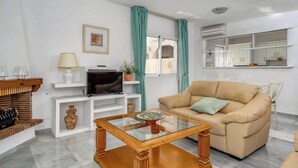 A living room with a beige sofa, a glass coffee table, a TV on a white stand, and a kitchen area with white cabinets and a window with green curtains. - Seaview Balcony by Costarentals - Ref 203 (Mijas)