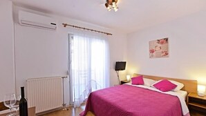 A bedroom with a bed, a radiator, a window with curtains, a wall-mounted TV, and a bedside table with a lamp.