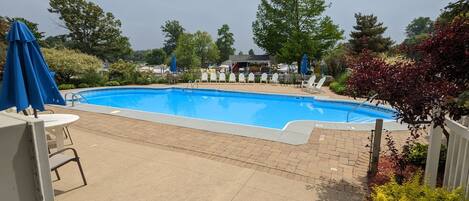 Una piscina rectangular con interior azul, rodeada por un solarium empedrado. Hay sillas de descanso blancas y una sombrilla azul cerca de la piscina.