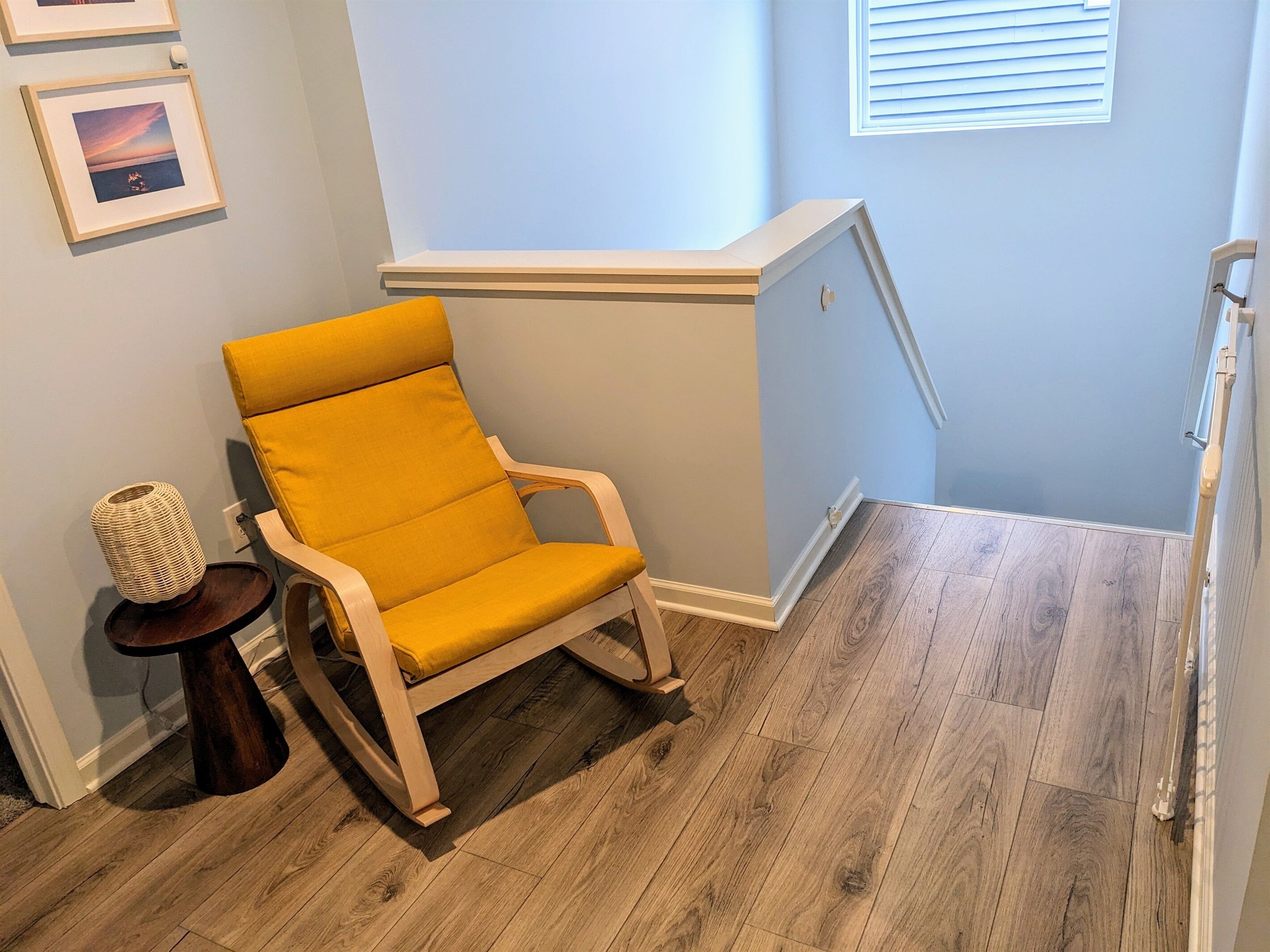 A corner room with a wooden floor, a yellow cushioned chair, a small round table with a woven basket, and a picture frame on the wall.