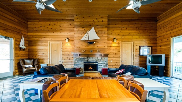A spacious living room with a wooden interior, a stone fireplace, leather sofas, a dining table, and ceiling fans.
