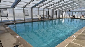 An indoor swimming pool with clear blue water, surrounded by a tiled deck and a glass ceiling structure.
