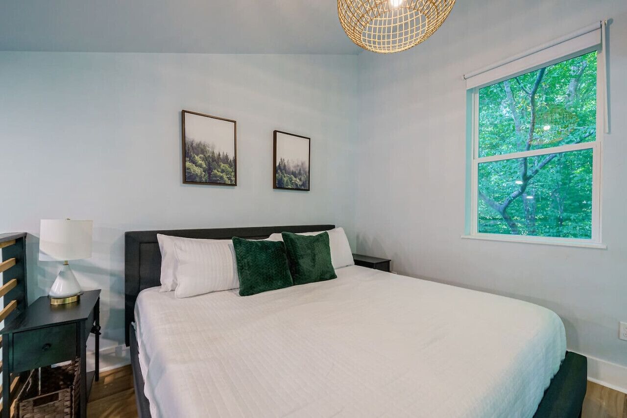 Un dormitorio con una cama, dos cuadros en la pared, una ventana con vistas a árboles y una lámpara sobre una mesita.