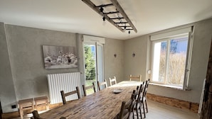 A spacious dining area with a long wooden table and chairs, large windows, and a ceiling light fixture. - Gîte à Saulxures sur Moselotte à 1 min du lac (Saulxures-sur-Moselotte)