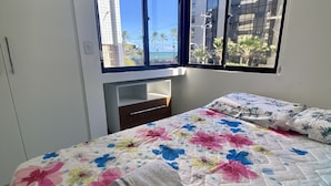 A hotel room with a bed, a nightstand, a window with a view of palm trees and buildings, and a ceiling vent. - Ametista 2 (Maceió)