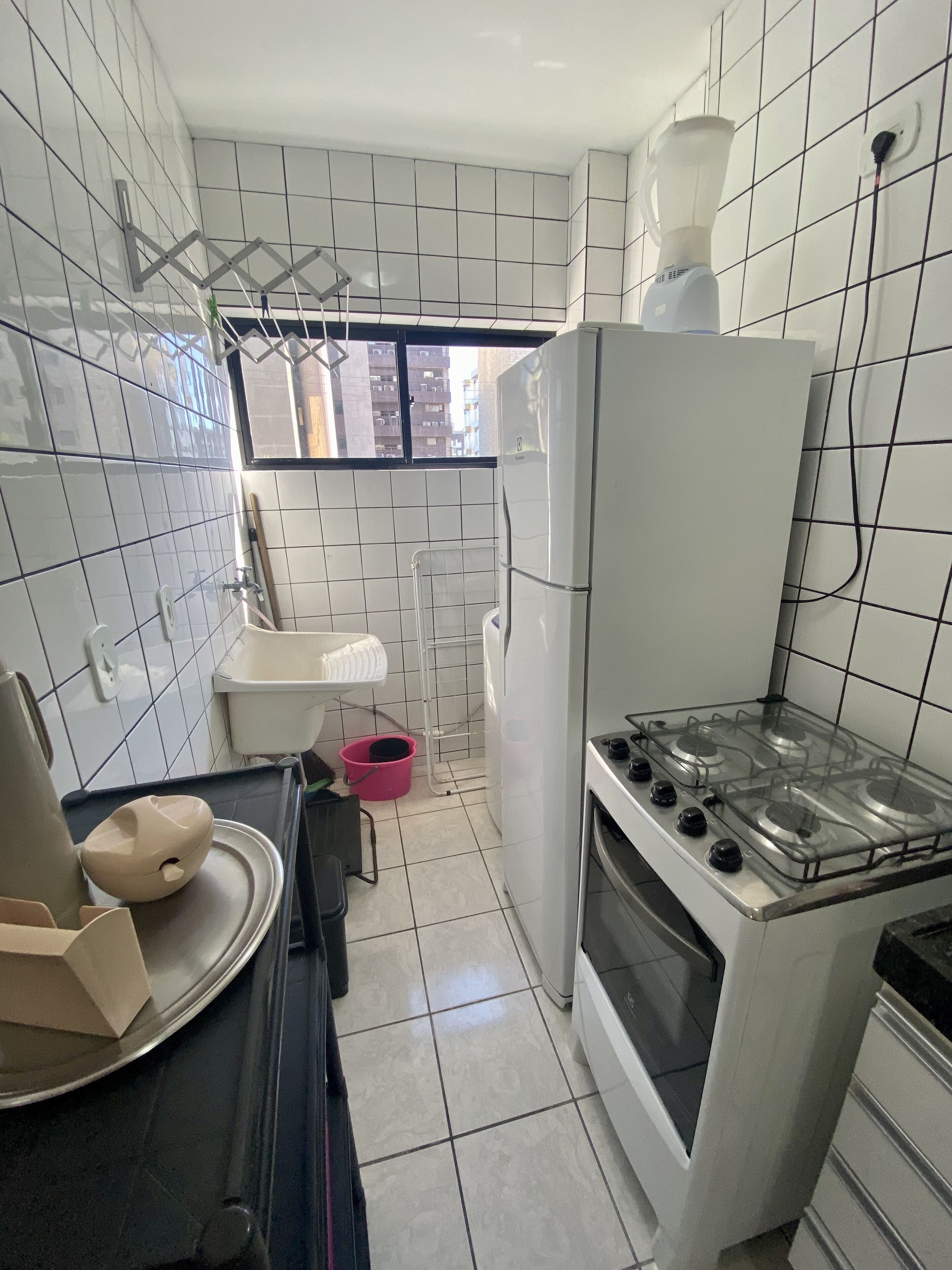 A compact kitchen with white tiled walls, a gas stove, a refrigerator, and a window with a view of buildings outside.