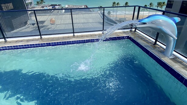 A swimming pool with a dolphin statue, a view of the ocean, and a building in the background. - Ametista 2 (Maceió)