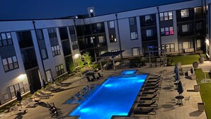 A swimming pool with lounge chairs and umbrellas, surrounded by a building with multiple balconies and a grassy area. - *LUXURIOUS 1BED/1BATH - KING BED* -POOL (Fishers)