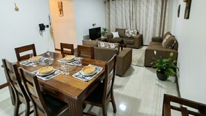A dining area with a wooden table set for four, surrounded by chairs. There is a sofa set in the background, a television on a stand, and a potted plant. - Breezeskyline with greenery view (Kiambu)