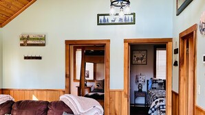 Interior rústico de uma cabana com teto de madeira, um sofá de couro com almofada que diz 'Bem-vindo à Cabana' e uma cama com cobertor xadrez.