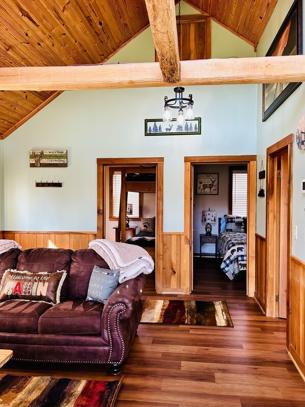 A rustic cabin interior with a wooden ceiling, a leather couch with a 'Welcome to the Cabin' pillow, and a bed with a checkered blanket. - Wilderness Retreat (Cadiz)