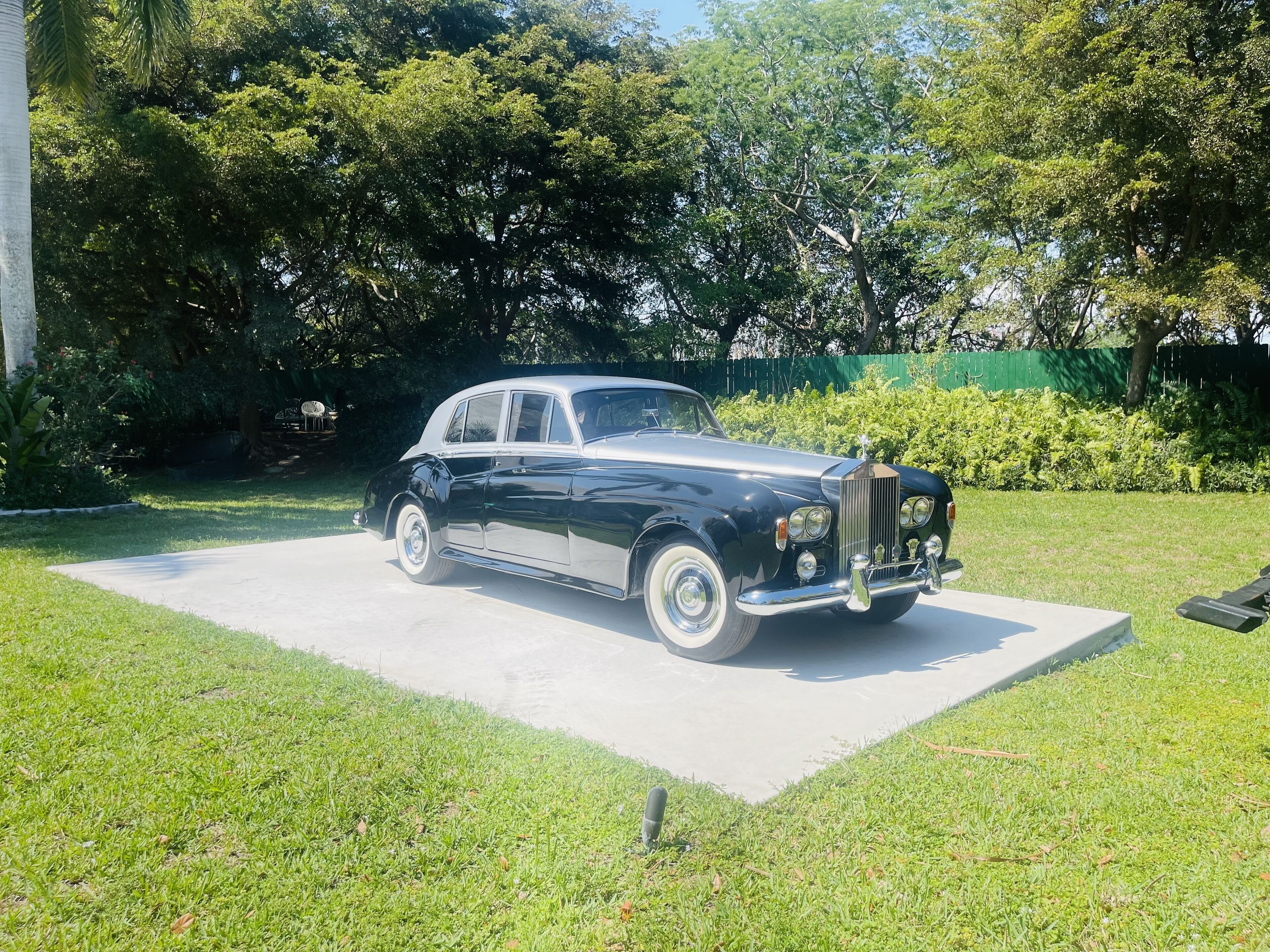 A classic black car parked on a concrete pad in a grassy area with trees and a fence in the background.