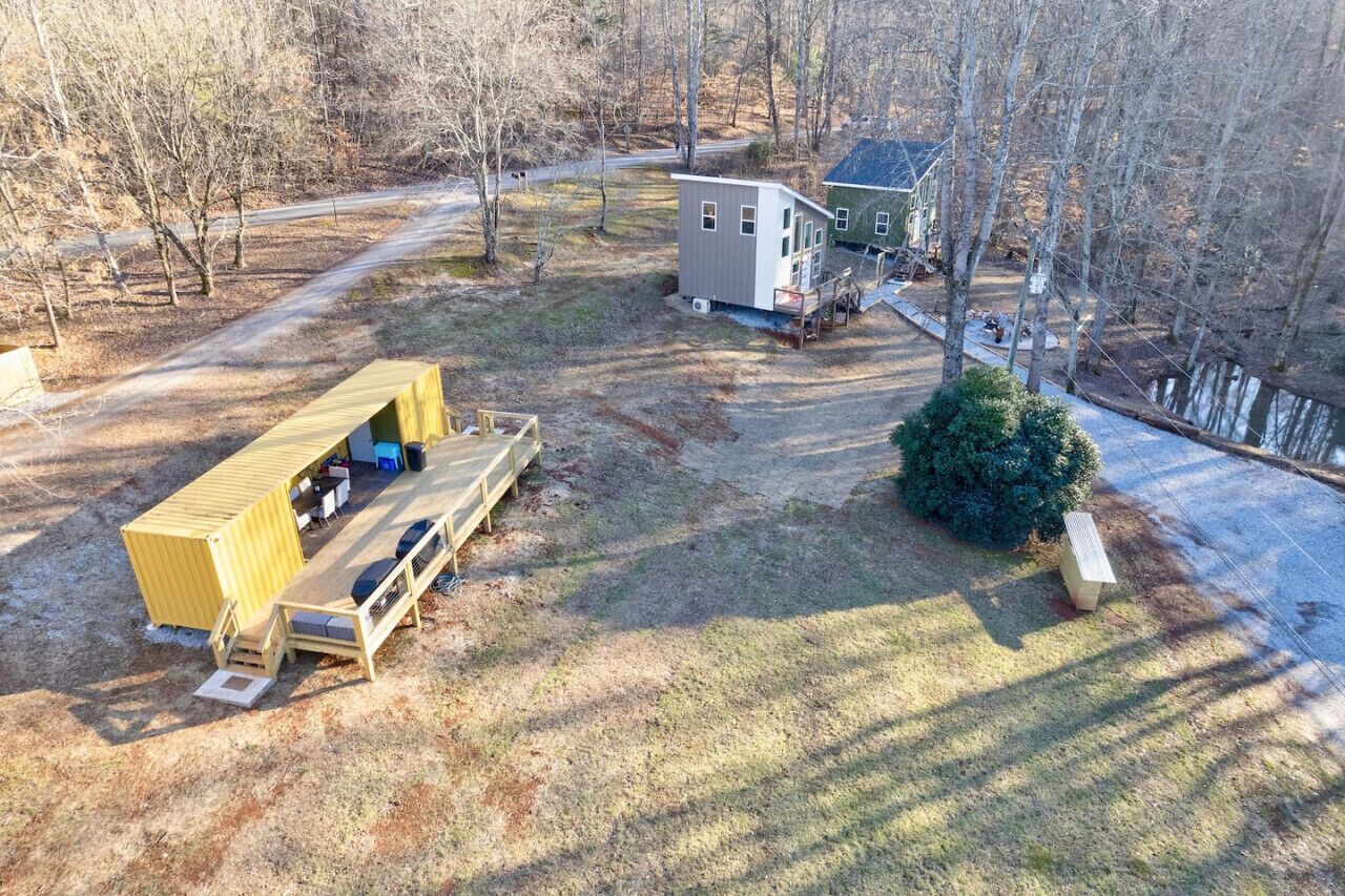 A rustic cabin with a deck and a small shed, surrounded by trees and a clear path.