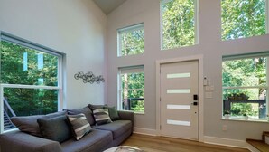 A modern living room with a grey sofa, a coffee table, and large windows offering a view of greenery outside.