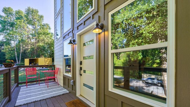 A wooden deck with a red chair and a small table, a door with a window, and a welcome mat.
