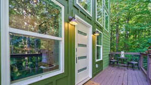 Une terrasse en bois de couleur verte, dotée d’une porte et de fenêtres en verre, d’un espace salon extérieur et offrant une vue sur les arbres environnants.