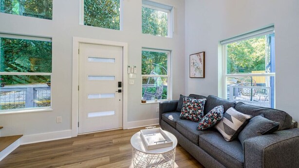A spacious living room with large windows, a grey sofa with patterned cushions, and a white door.