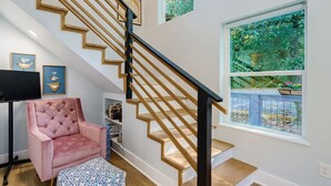 A staircase with wooden steps and black railings, a pink tufted armchair, and a large window with a view of greenery.