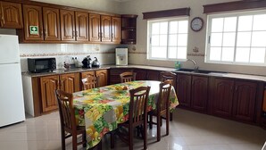 A kitchen with wooden cabinets, a dining table with chairs, a microwave, a refrigerator, and a window with a clock. - Green Tropical House ,Great view of the ocean (Ponta Delgada)