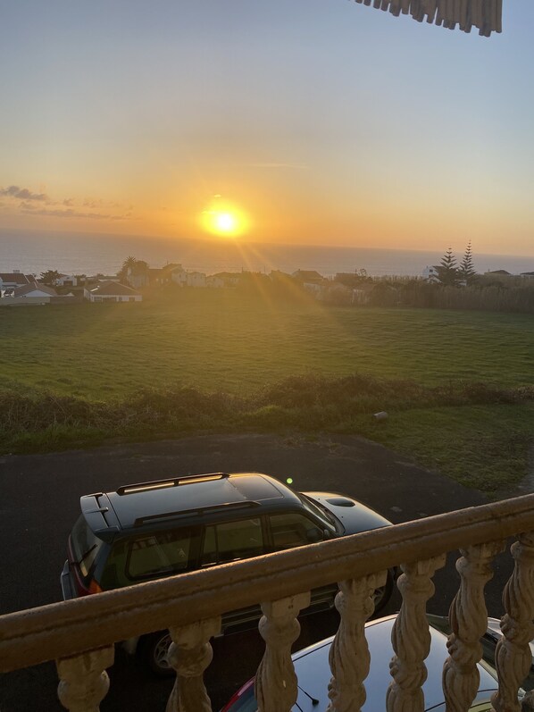 A sunset view from a balcony overlooking a parking area with a vehicle, grassy fields, and a distant shoreline. - Green Tropical House ,Great view of the ocean (Ponta Delgada)
