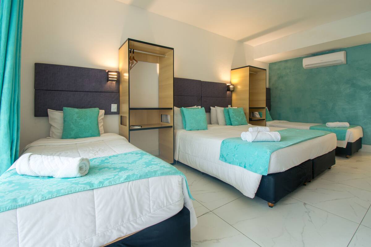 Une chambre d’hôtel avec trois lits, chacun doté d’une tête de lit, de draps blancs et d’éléments de décoration de couleur turquoise. Un grand miroir orne un des murs, et un appareil de climatisation est installé sur un autre.