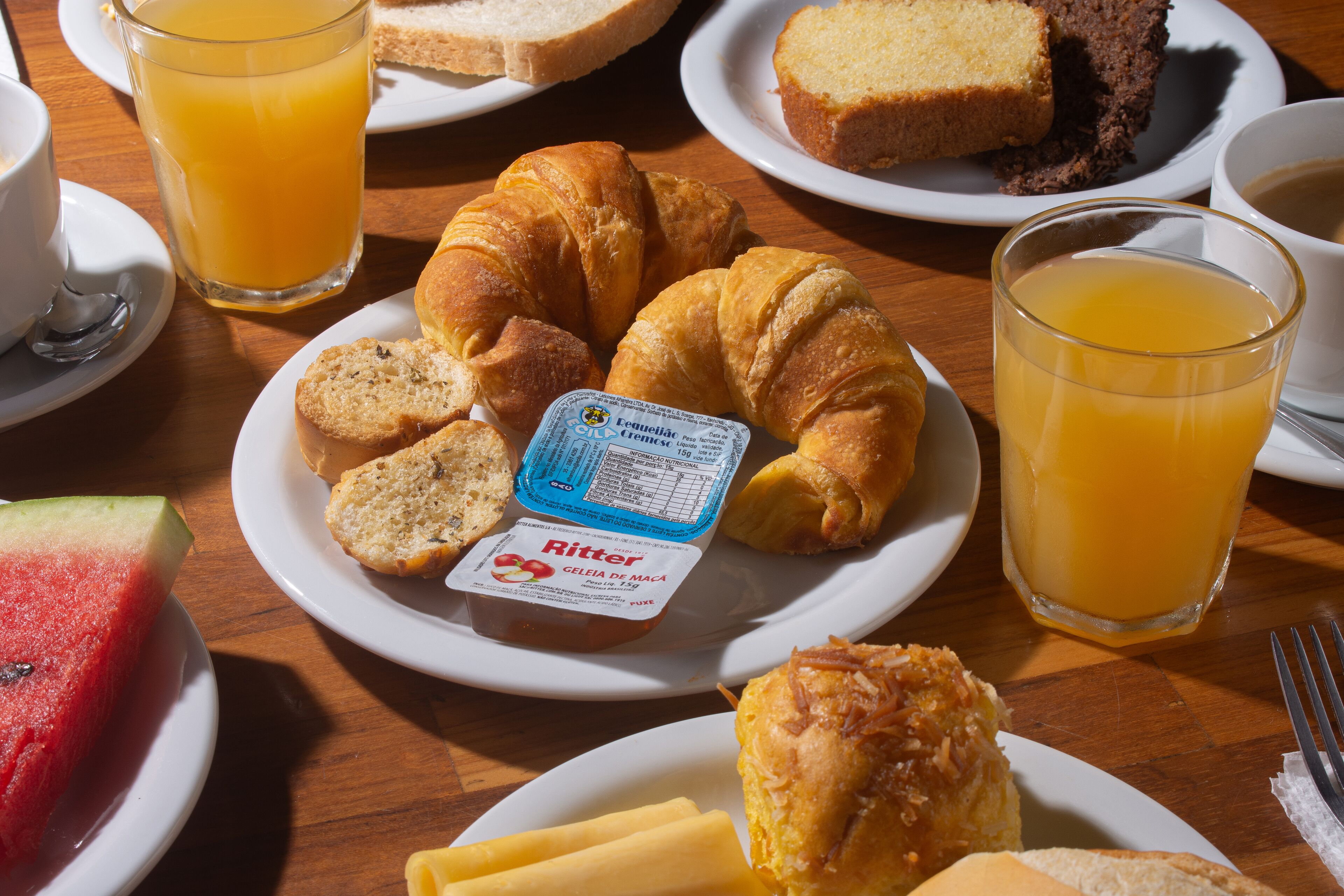 A breakfast setting with croissants, bread, a slice of watermelon, and a glass of orange juice on a wooden table.