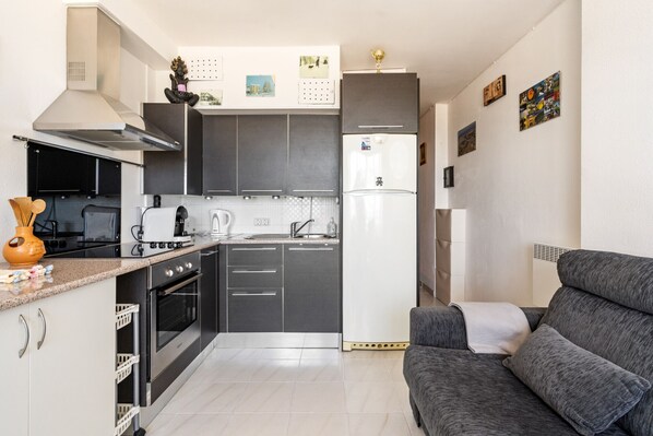 Una cocina compacta con armarios oscuros, un refrigerador blanco y un sofá gris en la sala.