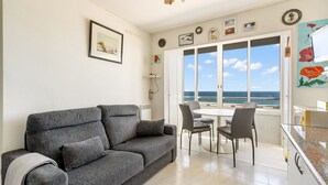 A living room with a grey sofa, a dining table with chairs, a kitchen area, and a large window with a view of the sea. - WELCS APARTAMENTO 053 EMP  Frente al Mar (Empuriabrava)