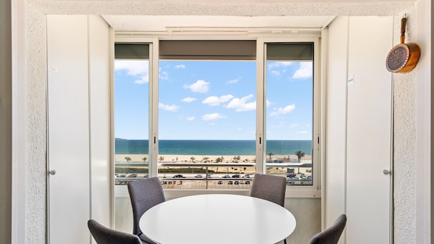 A room with a round table and four chairs, a view of the beach and ocean through large windows, and a door with a brass handle. - WELCS APARTAMENTO 053 EMP  Frente al Mar (Empuriabrava)