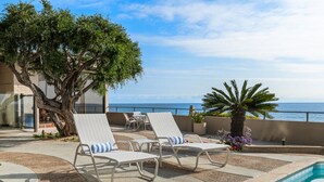 Zona de piscina con sillones de descanso, una palmera y vistas al mar.