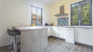Una cocina moderna con una isla central, muebles blancos, una campana extractora de acero inoxidable y un ventanal con vistas a una ladera.