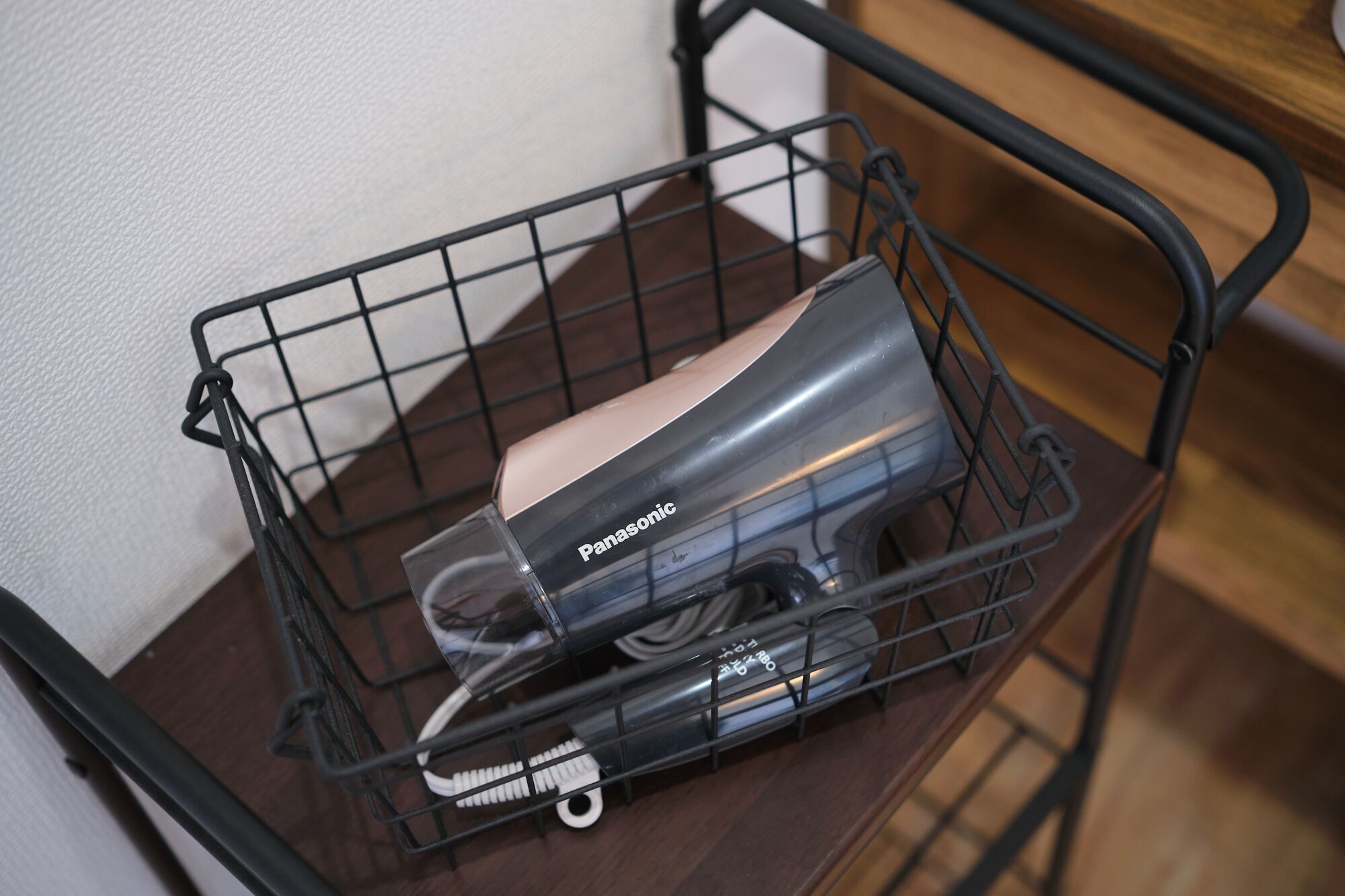 A black metal wire basket containing a Panasonic appliance, possibly a vacuum cleaner, on a wooden surface.