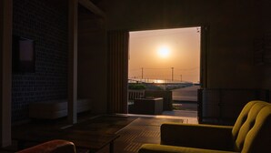 Une pièce offrant une vue sur le coucher de soleil à travers une porte ouverte, avec un fauteuil en osier et une petite table.