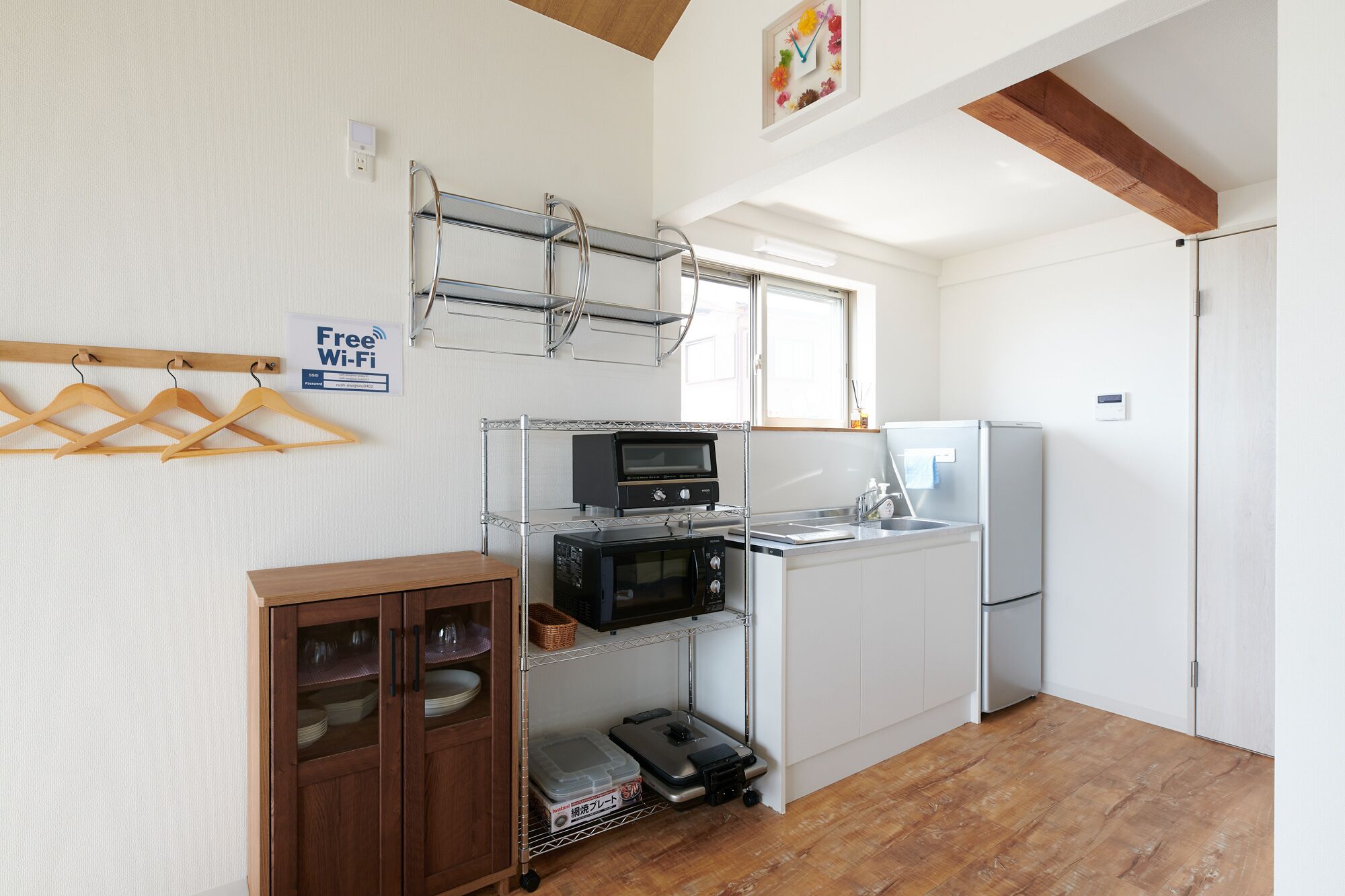 A compact kitchen with a built-in oven, microwave, and refrigerator. There is a wooden cabinet with glass doors and a metal rack with wooden hangers. A 'Free Wi-Fi' sign is visible on the wall.