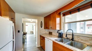 Una cocina con armarios de madera, un lavavajillas blanco, encimera de mármol y un ventanal con vistas al exterior.