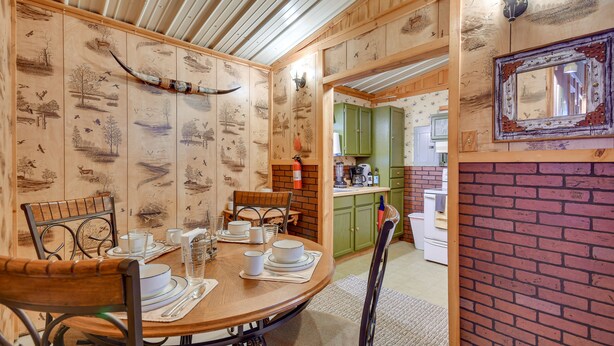 A dining area with a wooden table set for four, surrounded by chairs. A brick wall and a mirror are visible in the background. - Stargaze, Hike & Kayak: Cabin Near Copper Breaks! (Crowell)