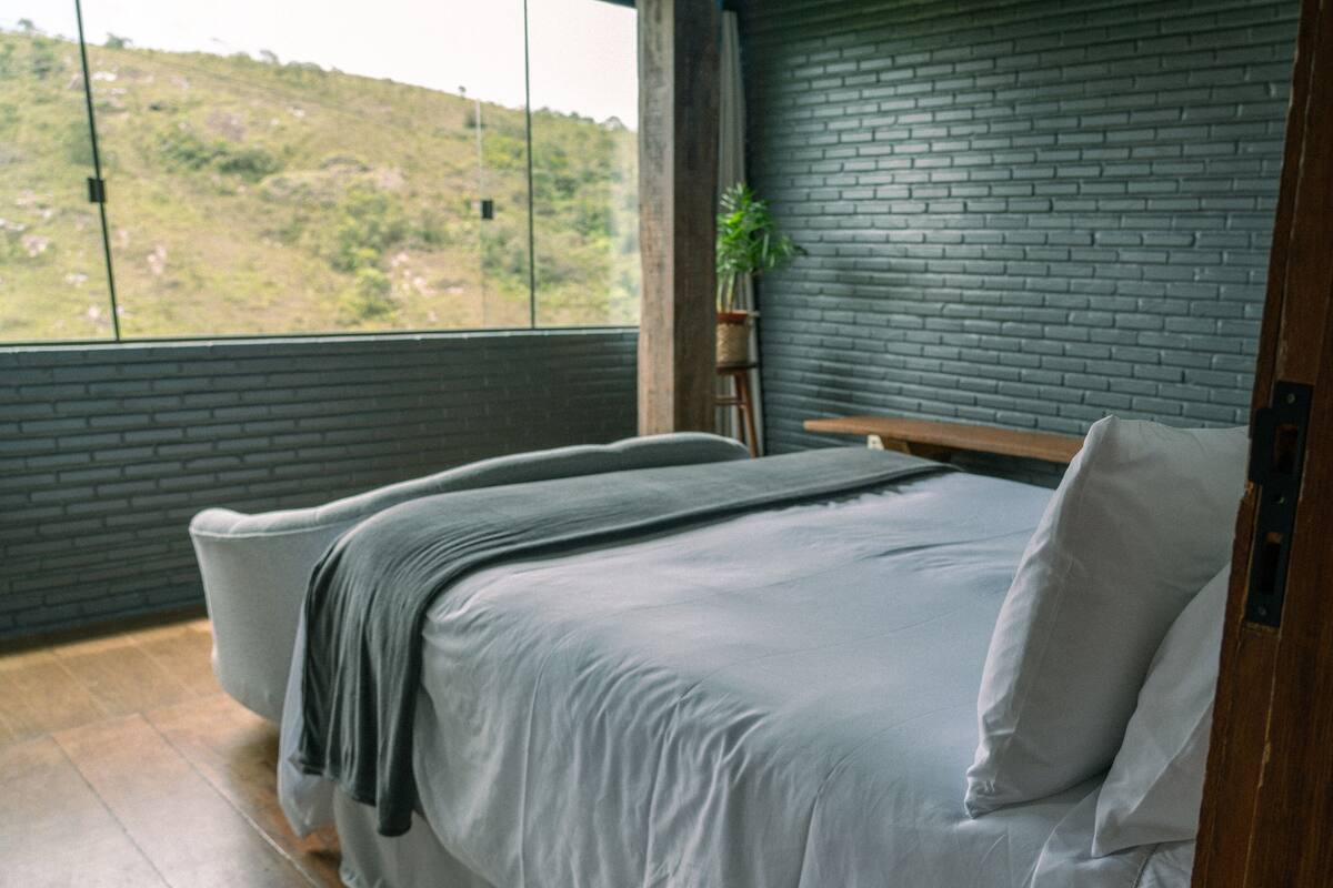 Un dormitorio moderno con un ventanal grande, pared de ladrillo, cama con ropa blanca y una planta en un estante.
