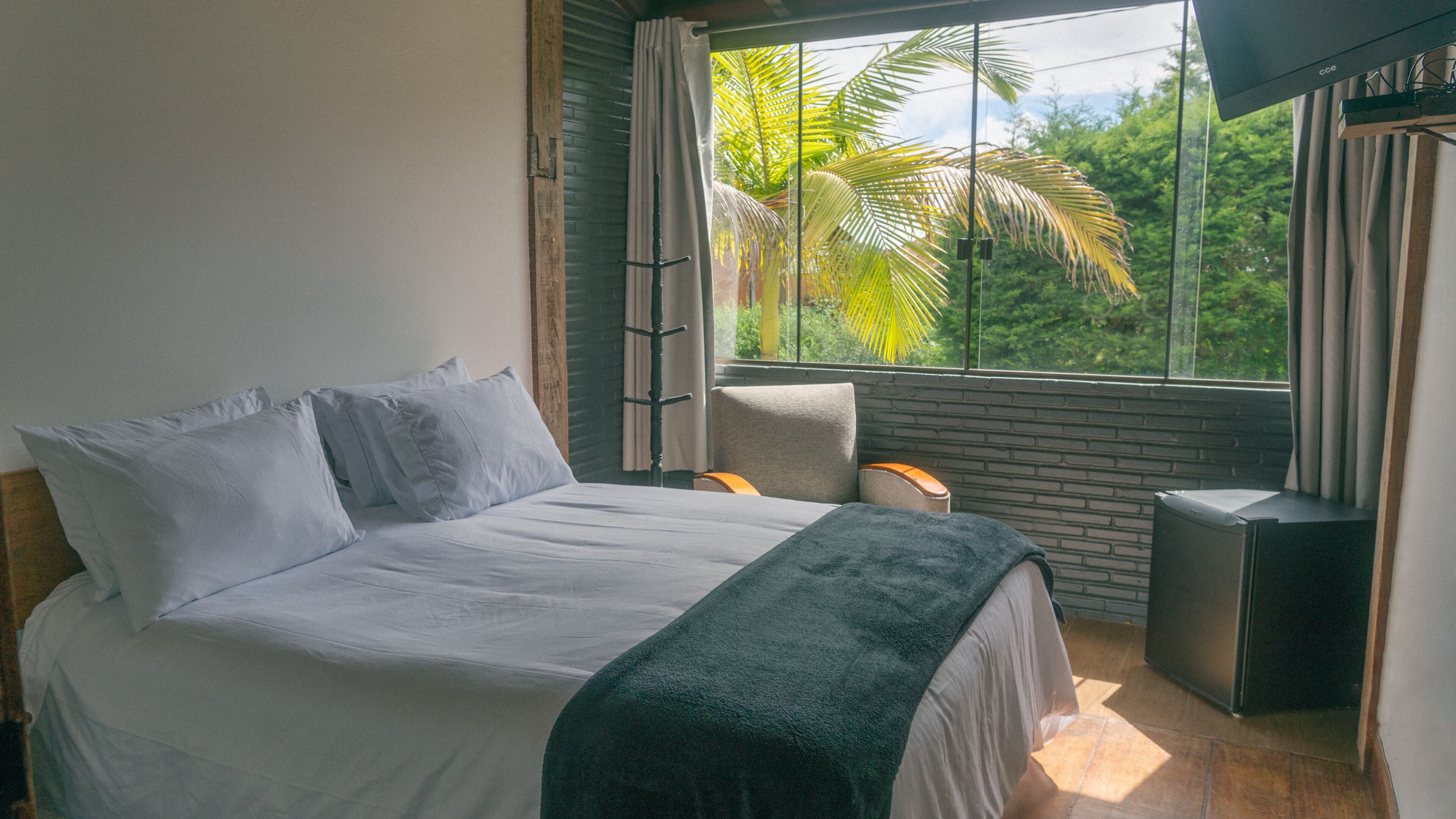 Un dormitorio con cama, una silla, televisión y ventana con vistas a follaje.
