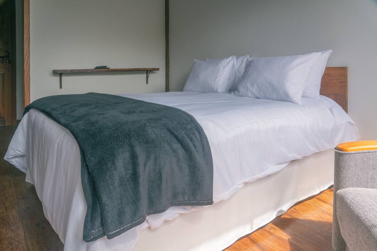Una cama bien tendida con sábanas blancas y un plaid verde oscuro, un cabecero de madera, y una pequeña estantería encima.