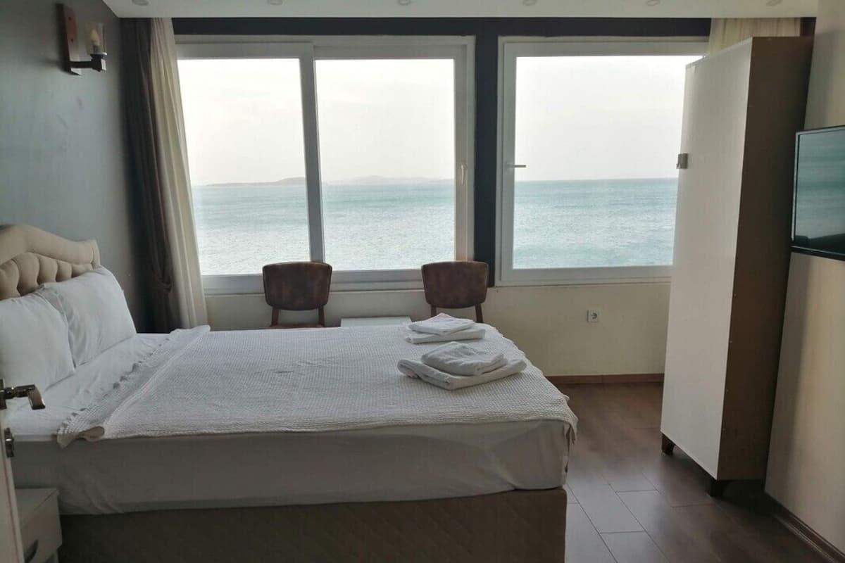 A hotel room with a bed, two chairs, a TV, and a large window overlooking the sea.