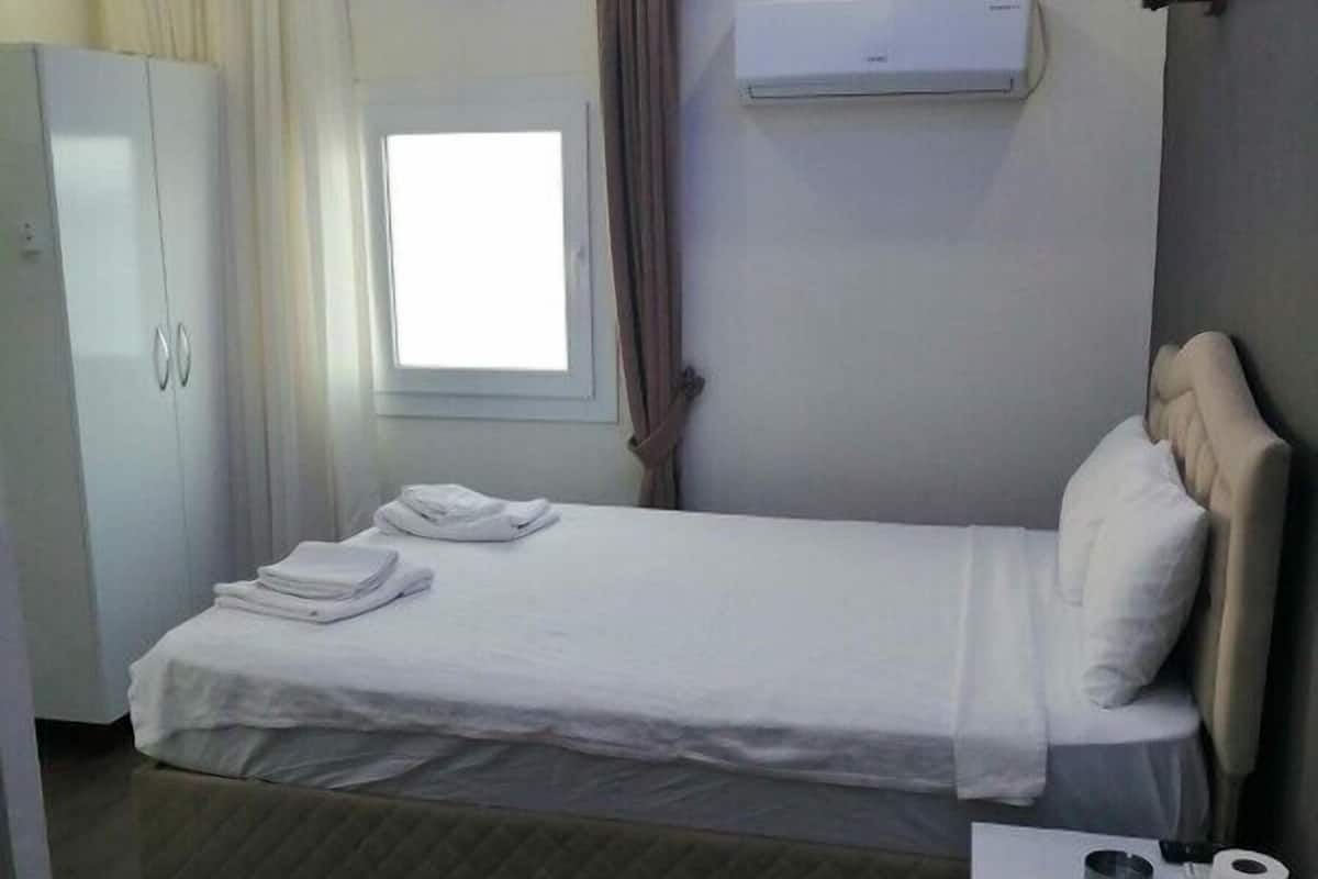 A hotel room with a bed, a small bedside table with a cup and a tissue, a wardrobe, and a window with curtains.