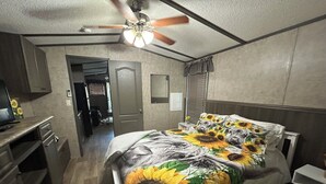 Ein Schlafzimmer mit einem mit Sonnenblumenmuster bezogenen Bett, einem Deckenventilator, einem Schreibtisch mit einem Computer und einer Tür zu einem anderen Raum.
